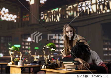 Person waking up african american girl in the university campus library, student falling asleep on her stack of borrowed books. Extremely tired and overwhelmed pupil tries to study all night. 125857908