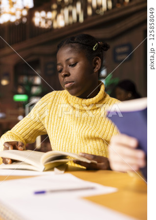 Young girl scholar reading literary works and taking notes in a library, focusing on curriculum based learning and academic success. Determined pupil enjoys mandatory books, school book club. 125858049