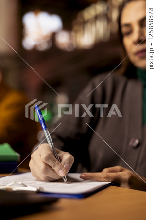 Adult learner transcribes information during a lecture in a university library, surrounded by academic textbooks and archived documents. Higher education personal goals on campus. Close up. 125858328