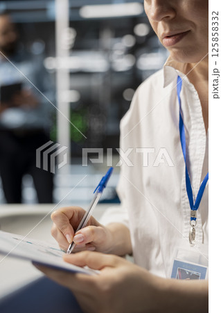 Woman in data center taking note on fixes to be made after running diagnostic scripts on computer. Server room admin writing repairing needed by supercomputers after evaluating performance metrics 125858332