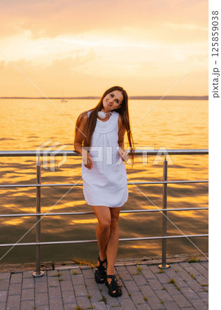 Young woman in white dress stands on pier by the water. The sky is orange and the water is calm Young woman in white dress stands on pier by the water. The sky is orange and the water is calm 125859038