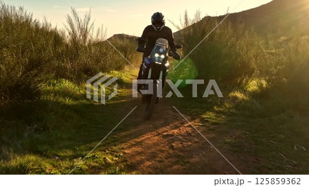Man, professional motorcyclist in full moto equipment riding crops enduro bike on mountain road at sunset. Concept of motosport, speed, hobby, journey, activity 125859362