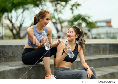 Female friends in athletic wear laughing and hydrating after physical activity in the park 125859643