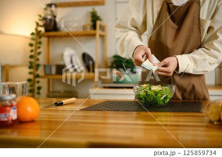 Close up of man in an apron pouring dressing onto fresh green salad in a modern kitchen Close up of man in an apron pouring dressing onto fresh green salad in a modern kitchen 125859734