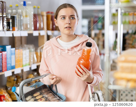 Girl customer in supermarket examines fruit juice, chooses drink in grocery store Girl customer in supermarket examines fruit juice, chooses drink in grocery store 125860620