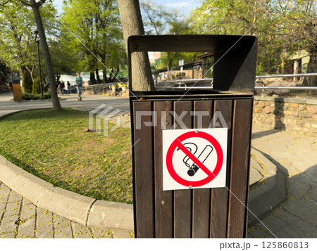 Close up of wooden outdoor trash bin with bold no smoking sign attached, placed in clean public park with trees on bright sunny spring day, promoting cleanliness 125860813