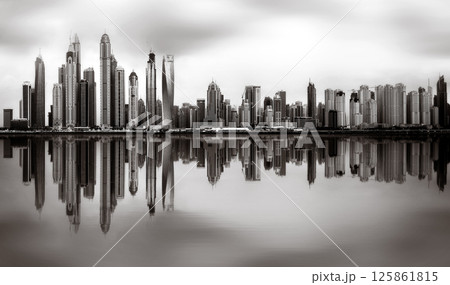 Monochrome city skyline with tall skyscrapers reflecting on calm water under a cloudy sky, Dubai Marina bay UAE 125861815