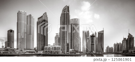 Monochrome city skyline with tall skyscrapers reflecting on calm water under a cloudy sky, Dubai Marina bay UAE 125861901