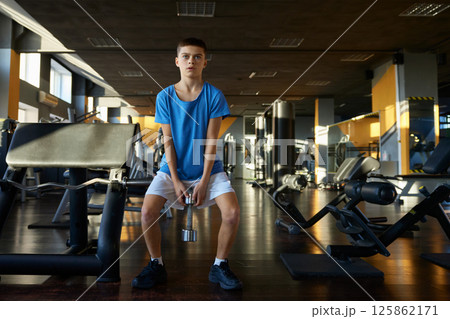 Athletic teenage boy squatting with dumbbells training over gym interior Athletic teenage boy squatting with dumbbells training over gym interior 125862171