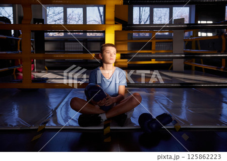 Teenage boy boxer resting after training inside boxing ring posing Teenage boy boxer resting after training inside boxing ring posing 125862223
