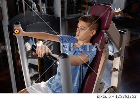 Teen boy doing sports exercise at machine for chest muscles pumping Teen boy doing sports exercise at machine for chest muscles pumping 125862252