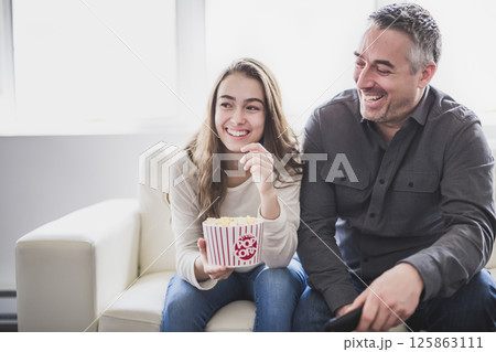 Portrait of a young man and daughter watching TV while eating popcorn on the sofa Portrait of a young man and daughter watching TV while eating popcorn on the sofa 125863111