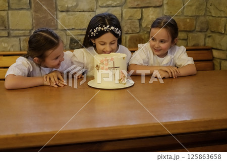Cheerful girls sit at the table with a luxurious cake. Three sisters, like little princesses, surround the dessert with curiosity Cheerful girls sit at the table with a luxurious cake. Three sisters, like little princesses, surround the dessert with curiosity 125863658