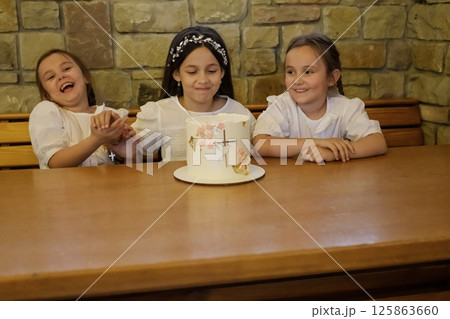 The girls near the festive cake look inspired and sincere. All three gently gaze at the decorated creamy cake. 125863660