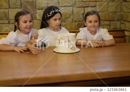 The girls are dressed festively and sit beside the elegant cake. The cake at the center of the table draws the gaze of the three little beauties. 125863661