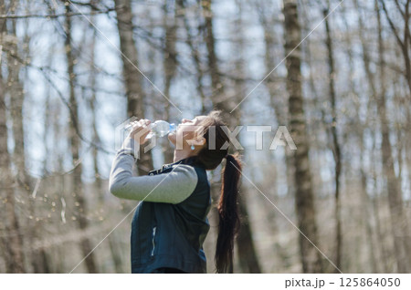 A black-haired woman drinking water from a plastic bottle in the forest. Beautiful girl in the park drinking water from a bottle. 125864050