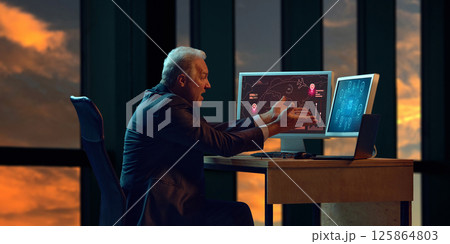 Elderly businessman sitting at office desk, gesturing with both hands in apparent shock while looking at two computer screens displaying complex data. 125864803