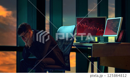 Businessman in suit sitting in office, two computer screens displaying downward trending graphs. 125864812