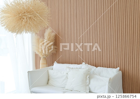 Soft white sofa with cushions and pampas grass in vase create cozy interior with natural wood panel wall and warm light for calm living space 125866754
