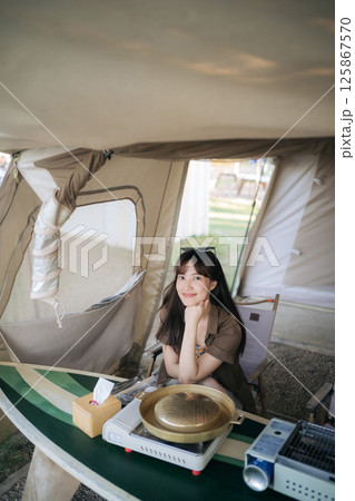 Southeast asian woman enjoys travel concept camping scene sitting inside tent with portable stove and grill on table smiling happily in natural light 125867570