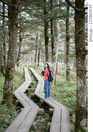 Southeast asian woman enjoys peaceful walk on wooden path through lush forest wearing casual clothes and backpack, surrounded by tall trees and green foliage Southeast asian woman enjoys peaceful walk on wooden path through lush forest wearing casual clothes and backpack, surrounded by tall trees and green foliage 125867637