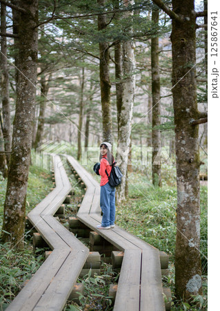 Southeast Asian woman enjoys peaceful walk on wooden path through lush forest wearing casual clothes and backpack with joyful expression on her face 125867641