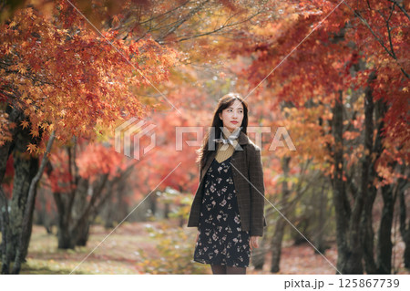 Southeast asian woman stands thoughtfully in forest filled with vibrant autumn leaves, wearing floral dress and jacket, surrounded by warm orange and red foliage Southeast asian woman stands thoughtfully in forest filled with vibrant autumn leaves, wearing floral dress and jacket, surrounded by warm orange and red foliage 125867739
