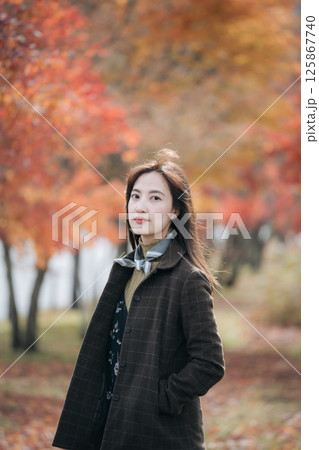 Southeast asian woman stands confidently in dark coat with autumn leaves in background creating warm and peaceful outdoor scene filled with natural beauty and calm expression 125867740