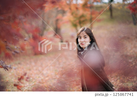 Southeast asian woman smiles gently while standing in serene autumn park surrounded by colorful red and orange leaves creating warm and peaceful atmosphere Southeast asian woman smiles gently while standing in serene autumn park surrounded by colorful red and orange leaves creating warm and peaceful atmosphere 125867741