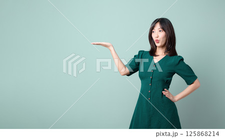 Southeast asian woman in green dress poses with advertising gesture showing something with her hand and playful expression on her face 125868214