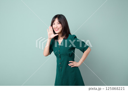 Cheerful southeast asian woman in green dress is advertising with bright smile and hand near her mouth on plain background Cheerful southeast asian woman in green dress is advertising with bright smile and hand near her mouth on plain background 125868221