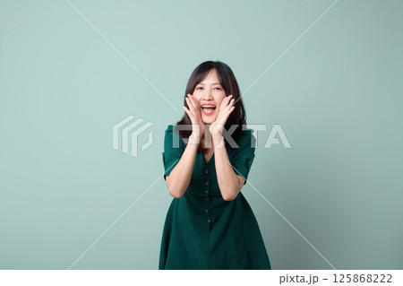 Southeast asian woman in green dress is happily shouting for advertising with excitement and joy on plain light green background Southeast asian woman in green dress is happily shouting for advertising with excitement and joy on plain light green background 125868222
