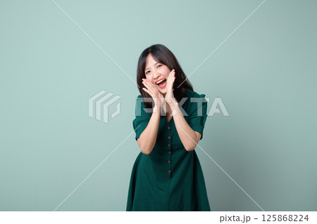 Cheerful southeast asian woman in green dress poses happily for advertising with hands near her face against plain light green background 125868224