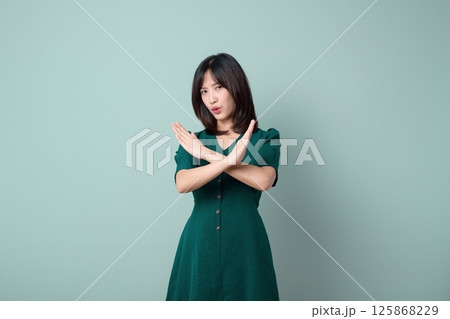 Southeast asian woman in green dress shows stop gesture with crossed arms expressing refusal for advertising or promotion in plain background Southeast asian woman in green dress shows stop gesture with crossed arms expressing refusal for advertising or promotion in plain background 125868229