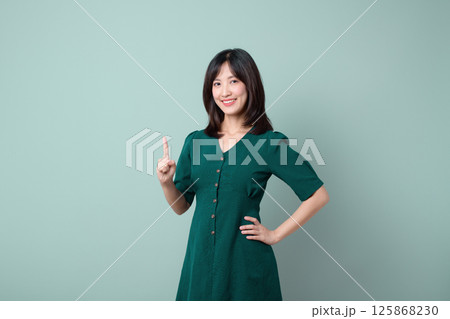Southeast asian woman in green dress is smiling confidently while advertising with raised finger against plain light green background Southeast asian woman in green dress is smiling confidently while advertising with raised finger against plain light green background 125868230