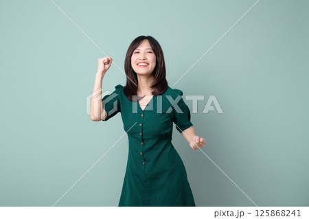 Southeast asian woman in green dress expressing joy and confidence for advertising with bright smile and raised fist on plain background 125868241
