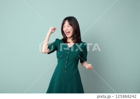 Happy southeast asian woman celebrating with closed eyes and fist raised in joy wearing green dress for advertising concept Happy southeast asian woman celebrating with closed eyes and fist raised in joy wearing green dress for advertising concept 125868242