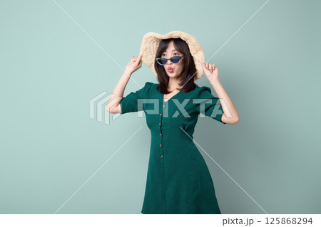 Southeast asian woman wearing sunglasses and hat poses playfully for advertising in green dress against light blue background 125868294