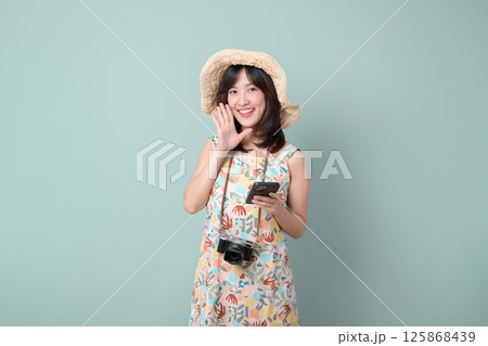 Southeast asian woman smiling happily while using mobile phone and internet application wearing floral dress and straw hat with camera hanging around her neck 125868439