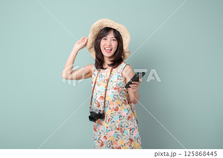 Southeast asian woman joyfully uses mobile phone to explore internet application while wearing floral dress and straw hat with camera hanging around her neck 125868445