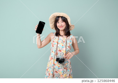 Southeast asian woman wearing floral dress and straw hat happily shows her phone screen while holding camera around her neck, enjoying online shopping and travel moments 125868446