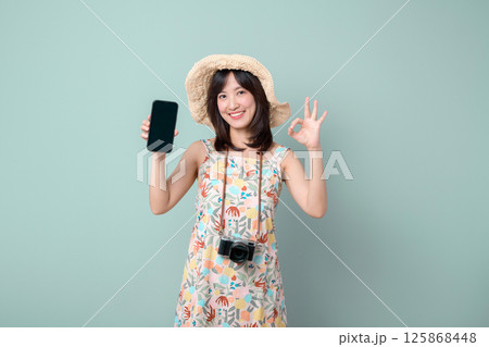 Southeast asian woman wearing floral dress and straw hat happily shows smartphone while making okay gesture symbolizing online shopping success and enjoyment Southeast asian woman wearing floral dress and straw hat happily shows smartphone while making okay gesture symbolizing online shopping success and enjoyment 125868448