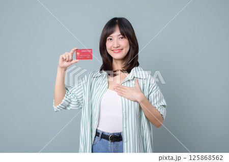 Southeast asian woman is happily shopping online holding credit card and smiling confidently while standing against plain background in casual clothes Southeast asian woman is happily shopping online holding credit card and smiling confidently while standing against plain background in casual clothes 125868562