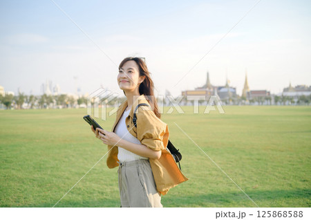 Southeast asian woman enjoys travel concept outdoors holding smartphone in large green field with historic buildings in background under clear sky Southeast asian woman enjoys travel concept outdoors holding smartphone in large green field with historic buildings in background under clear sky 125868588