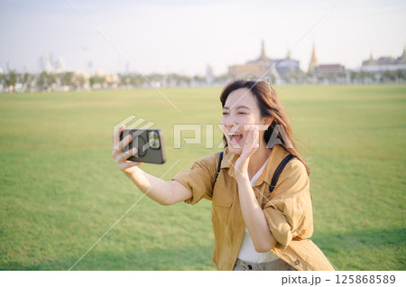 Southeast asian woman happily taking selfie with smartphone in large green park during travel concept, enjoying sunny day and capturing joyful moments outdoors Southeast asian woman happily taking selfie with smartphone in large green park during travel concept, enjoying sunny day and capturing joyful moments outdoors 125868589