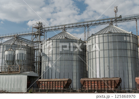 Large metallic silos feature prominently at a grain storage facility amidst a cloudy sky and scattered fields 125868970