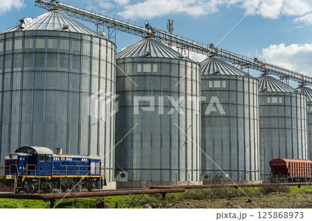 A blue locomotive rests beside a row of towering metal silos in a rural area, showcasing industrial agriculture and machinery A blue locomotive rests beside a row of towering metal silos in a rural area, showcasing industrial agriculture and machinery 125868973