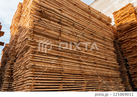 Wooden planks are neatly stacked in a lumber yard, reflecting sunlight on a clear day, next to a construction warehouse 125869011