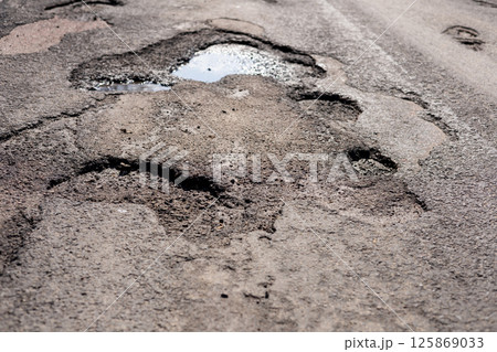 Potholes and rough patches cover the asphalt road with rainwater pooling in depressions, indicating poor maintenance and weather effects 125869033