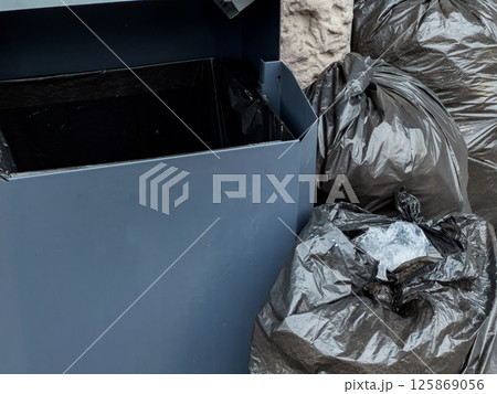 Piles of black garbage bags are placed next to a half-empty trash bin in an urban setting under natural light Piles of black garbage bags are placed next to a half-empty trash bin in an urban setting under natural light 125869056